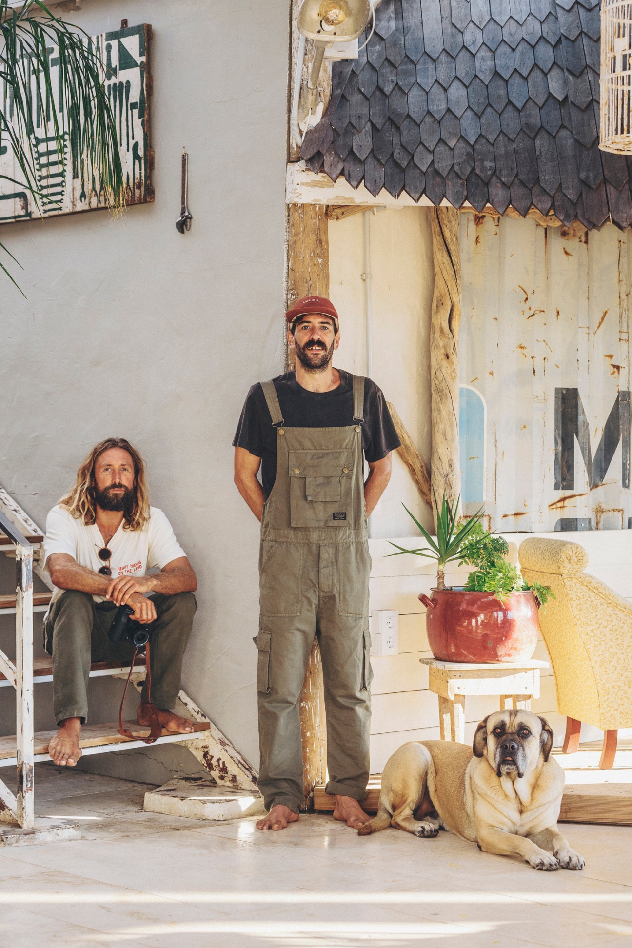 Two men stand in front of a weathered coastal building with a dog; one seated, one standing in work overalls.
