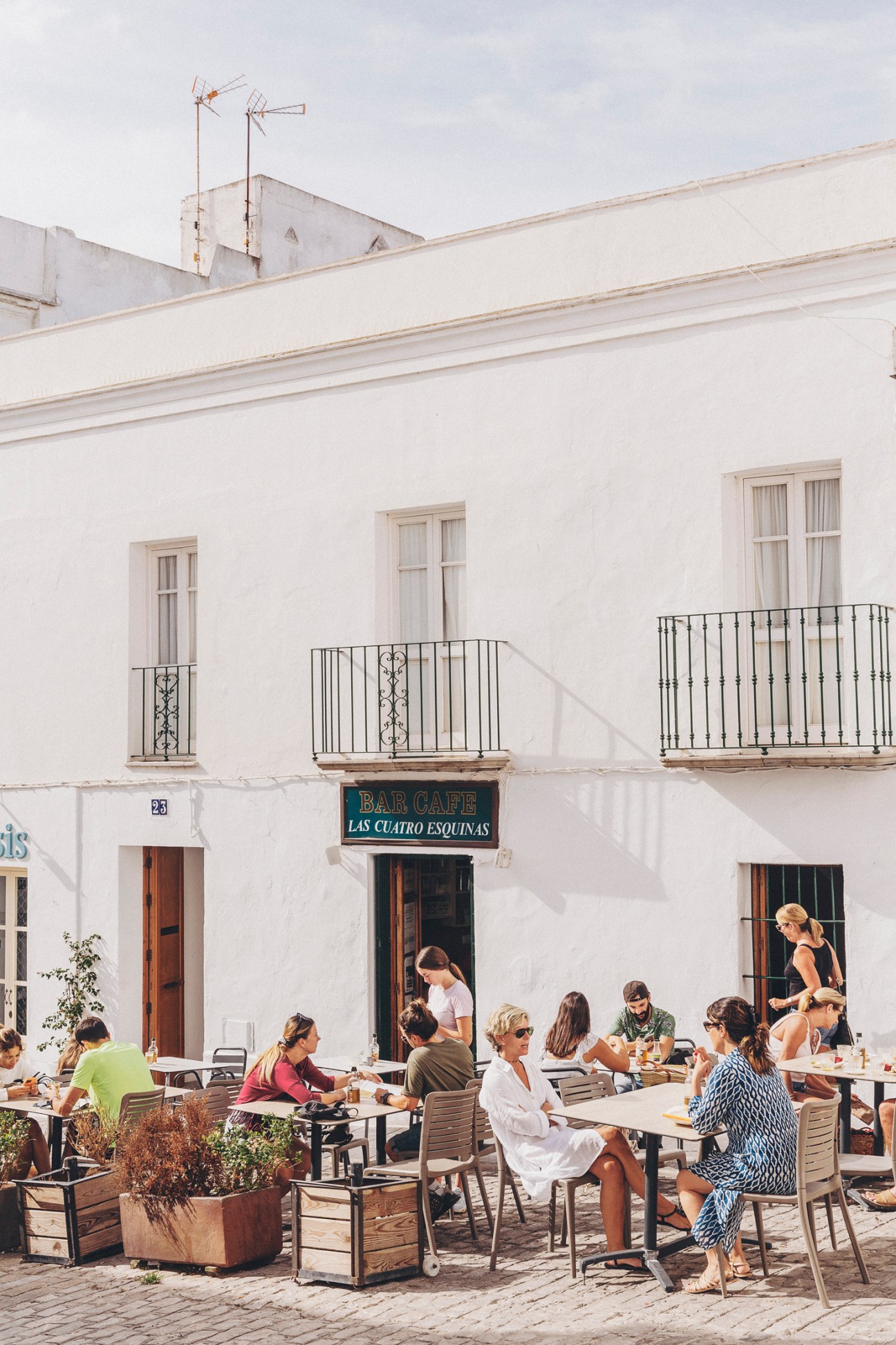 Outdoor café terrace with white-washed building facade, diners seated at tables, and black iron balconies above.