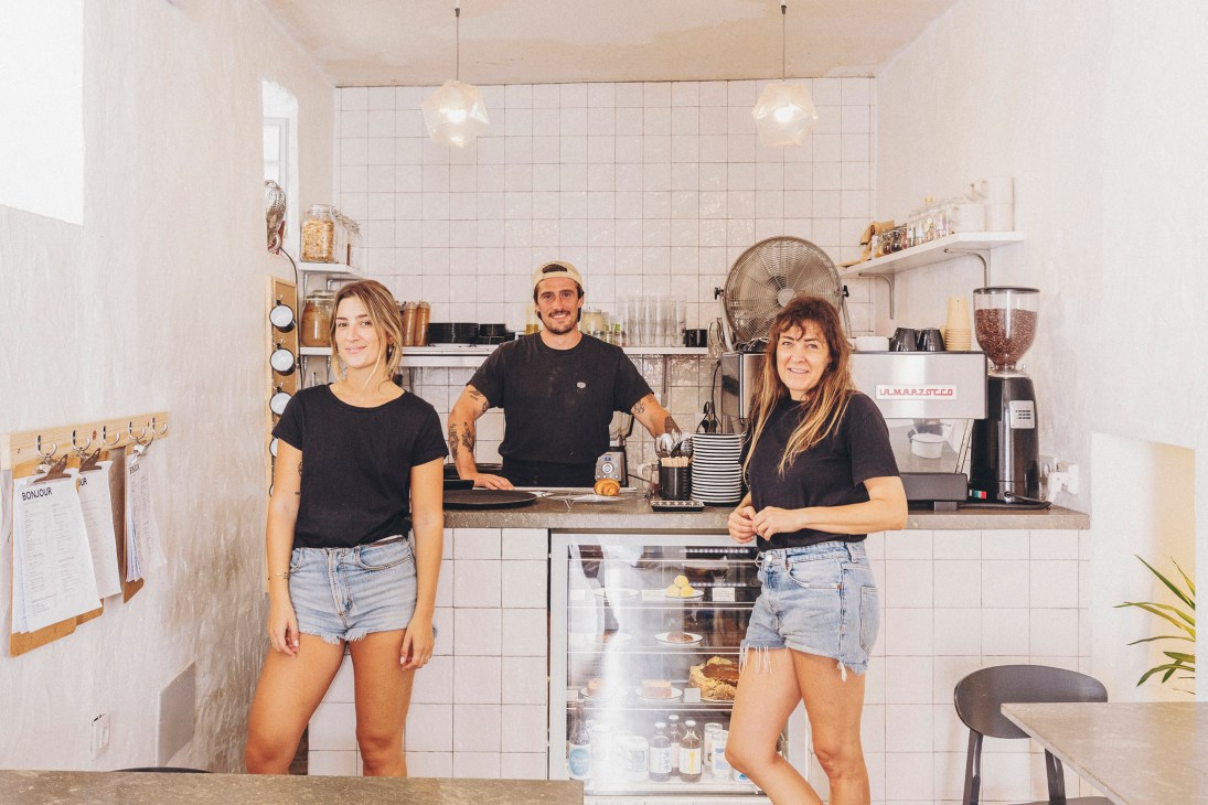 Three staff members stand behind the counter at Bonjour Café in Tarifa, a minimalist white-tiled café with pendant lights and shelving.