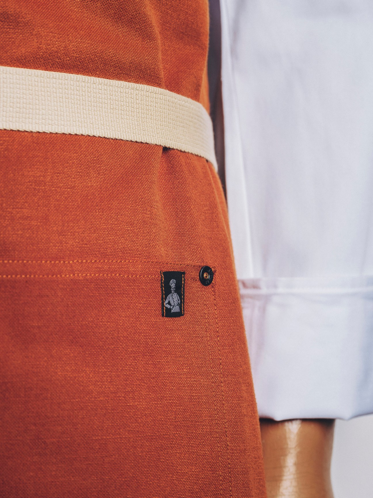Close-up of orange fabric apron with cream waistband showing detailed stitching and small black label with chef logo