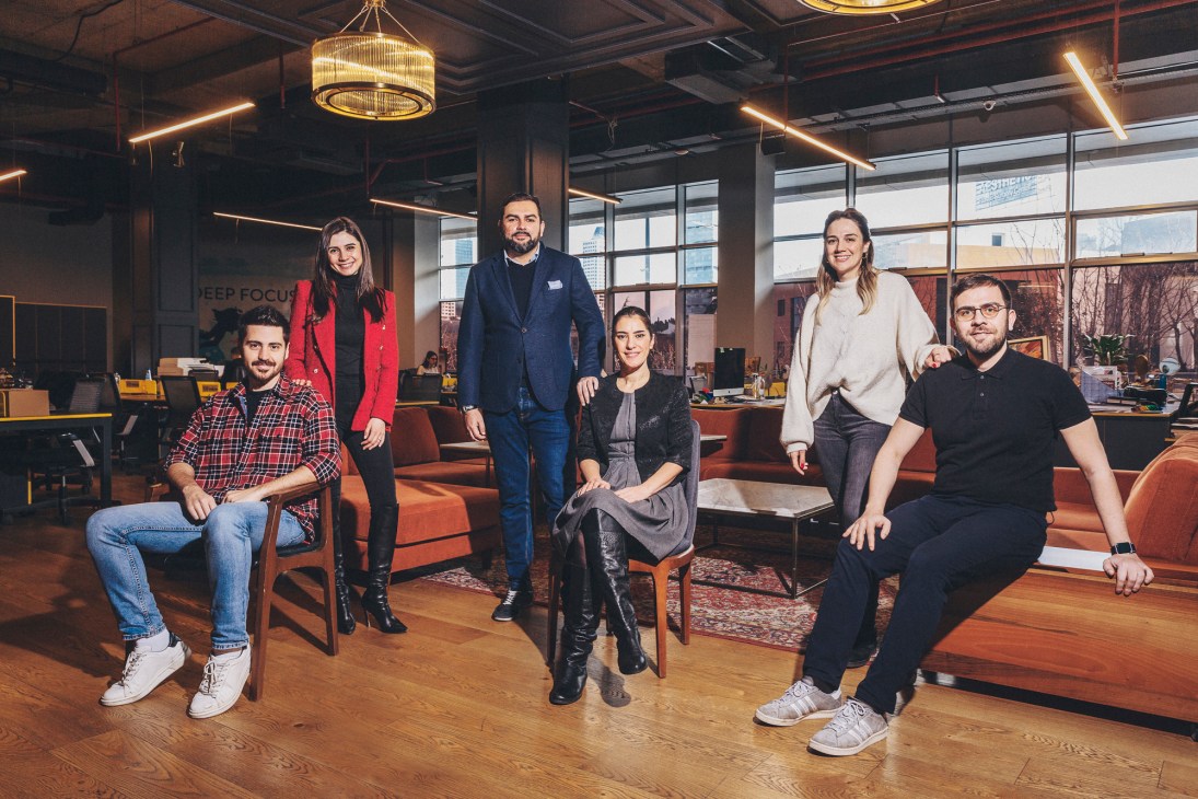 Group of six professionals posing together in a modern office with exposed ceiling and large windows