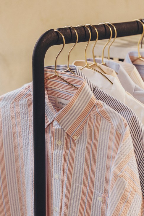 Striped shirts hanging on a brass rail with black rod, showing Unfeigned brand label.