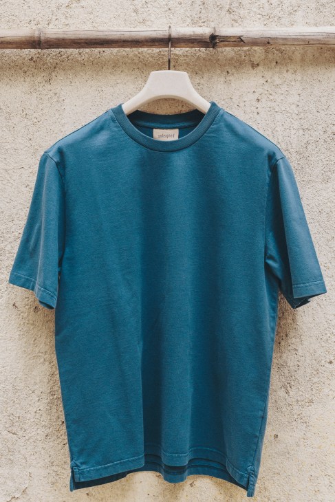 Blue Unfeigned Mediterranea T-shirt on white hanger against concrete wall.