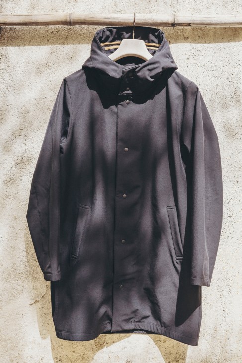 Unfeigned AW21 charcoal grey trench coat with hood hanging on a wooden rail against a weathered concrete wall.