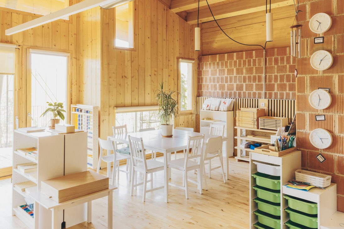 Wooden classroom interior with exposed ceiling beams, white furniture, clocks on wall, and educational storage