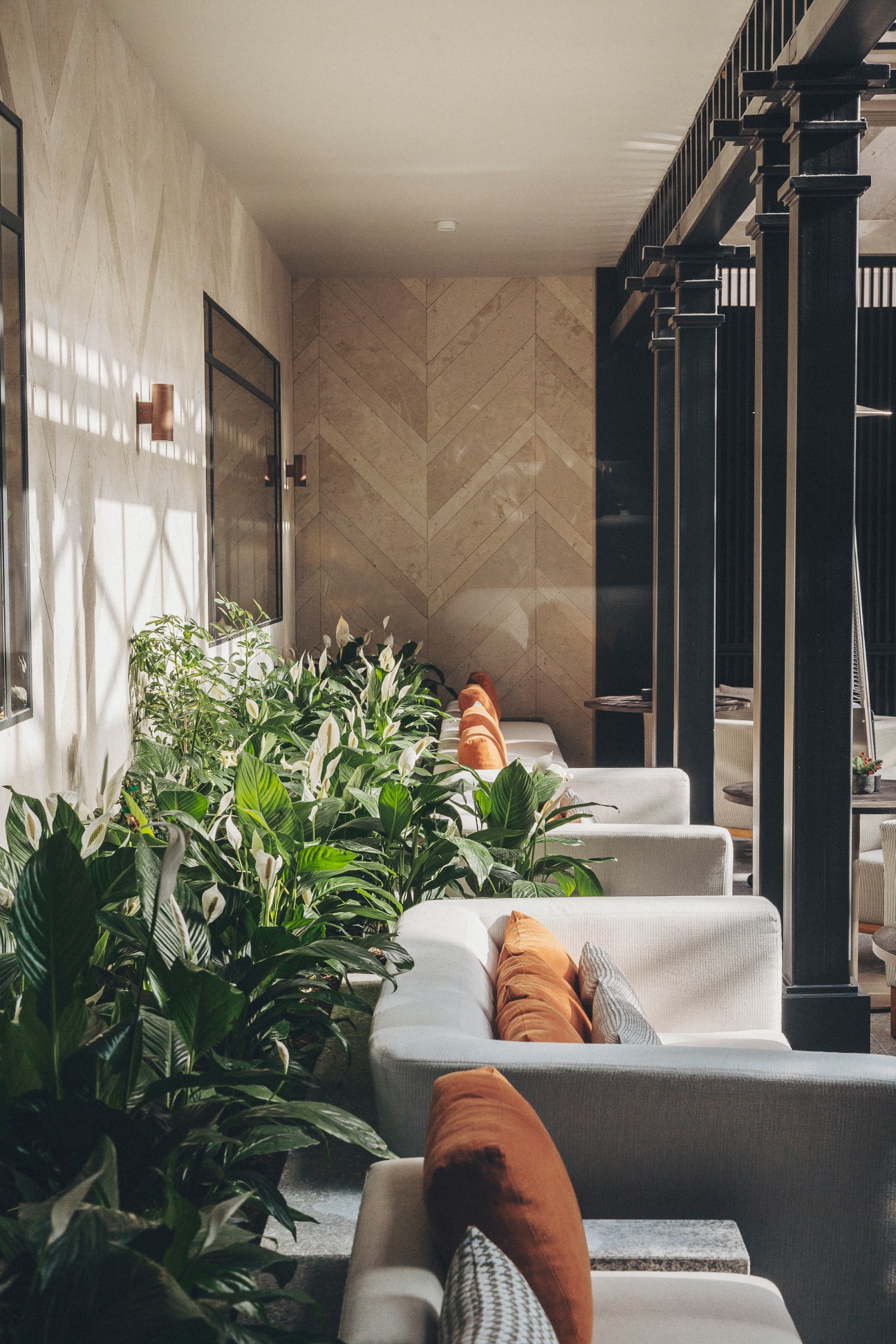 Modern hotel lobby seating area with white armchairs, orange cushions, lush green plants and dark structural columns