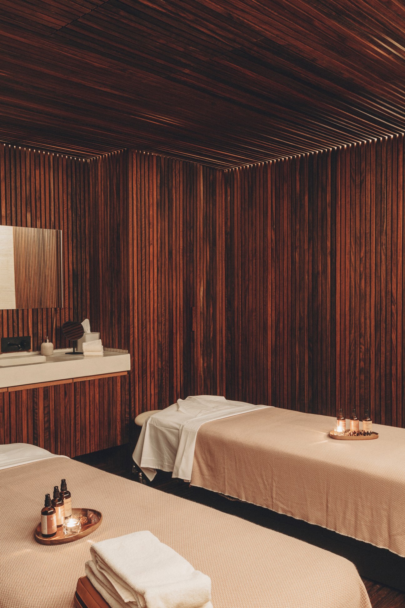 Spa treatment room with wooden slat walls, two massage tables with cream linens, and wellness accessories