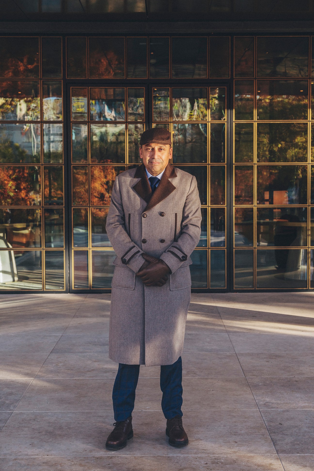 Dapper doorman in long grey coat and cap standing in front of glass-windowed hotel building
