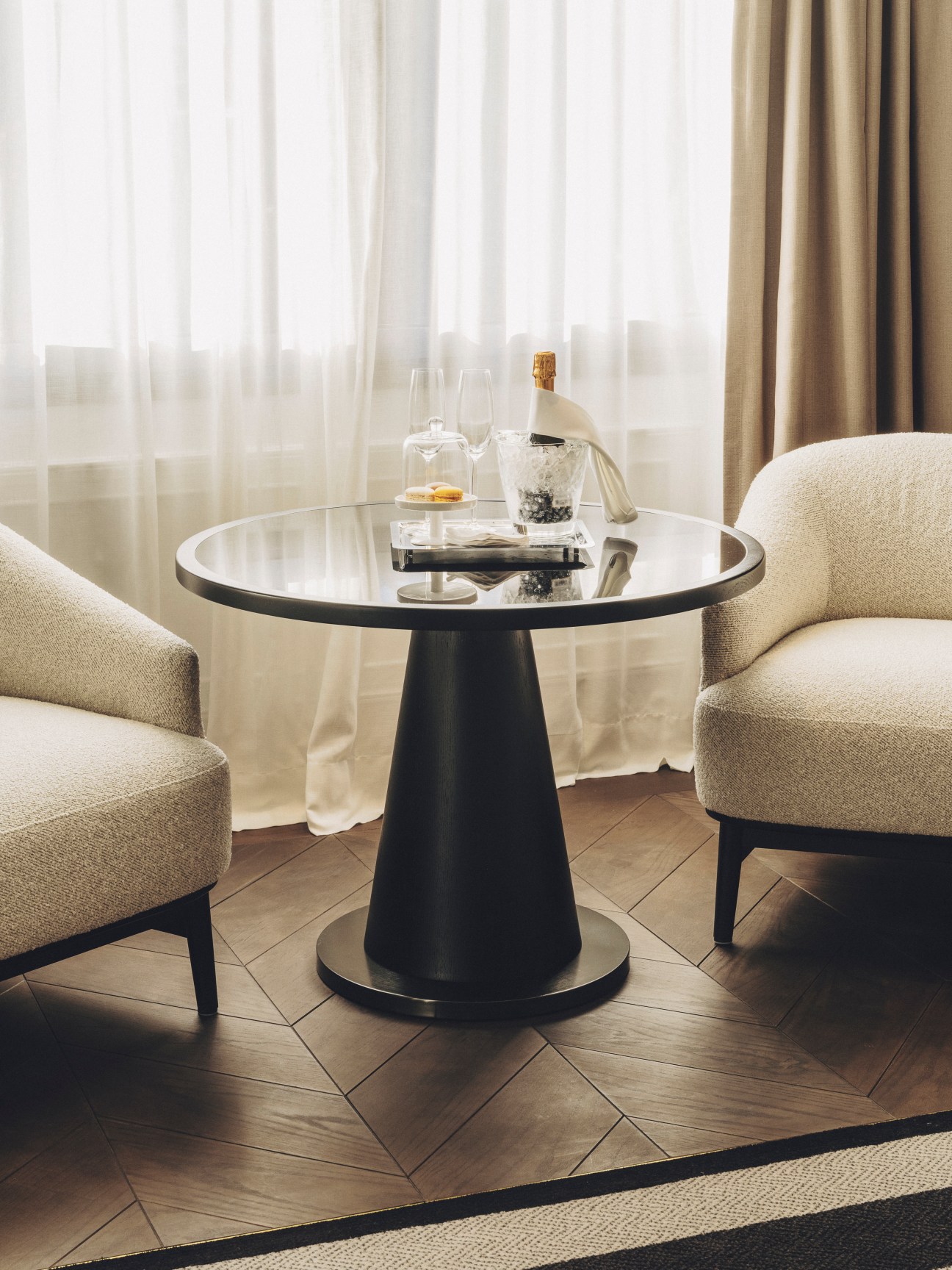 Elegant hotel seating area with round glass table, champagne bottle, wine glasses and neutral armchairs by window