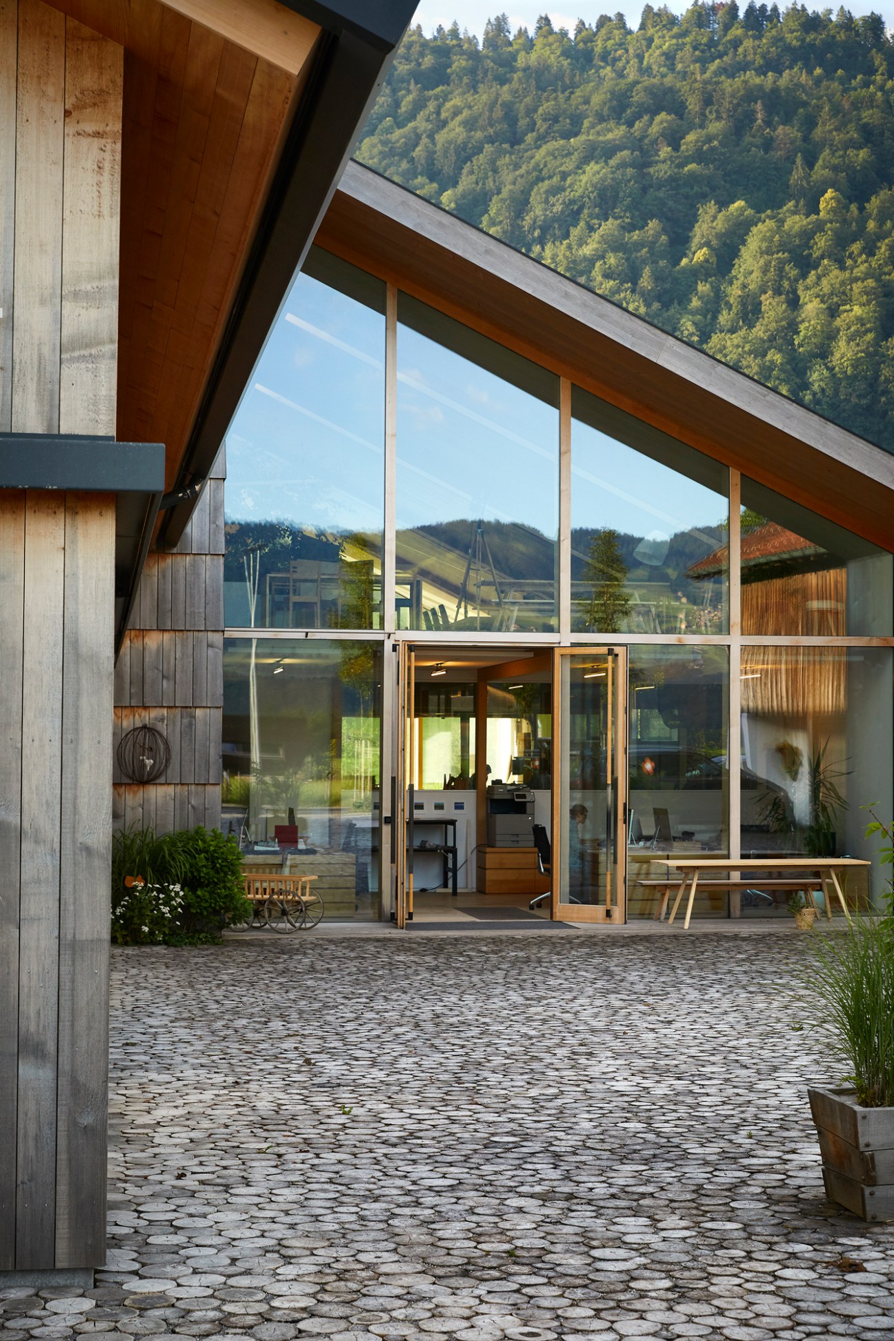 Modern timber and glass workshop building with large windows and cobblestone courtyard surrounded by forested hills