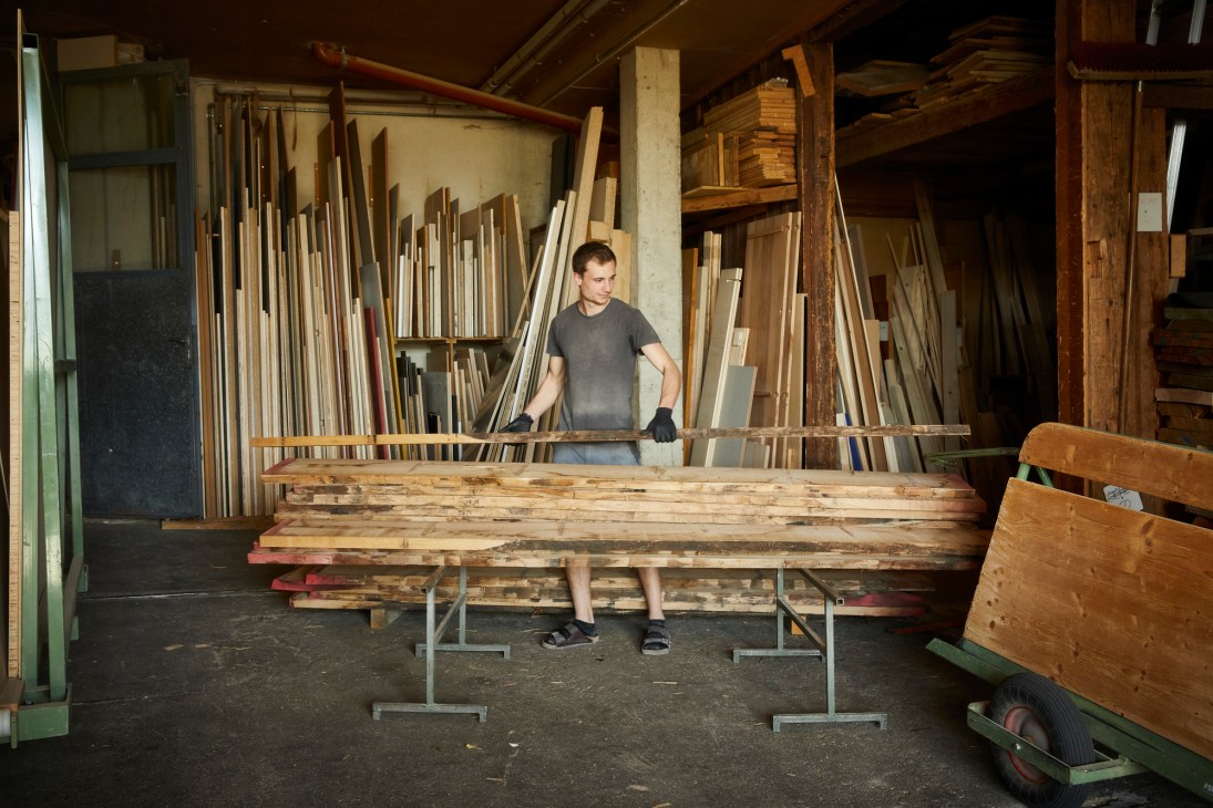 Man in gray t-shirt standing behind stacks of wooden lumber planks organized on metal supports in workshop