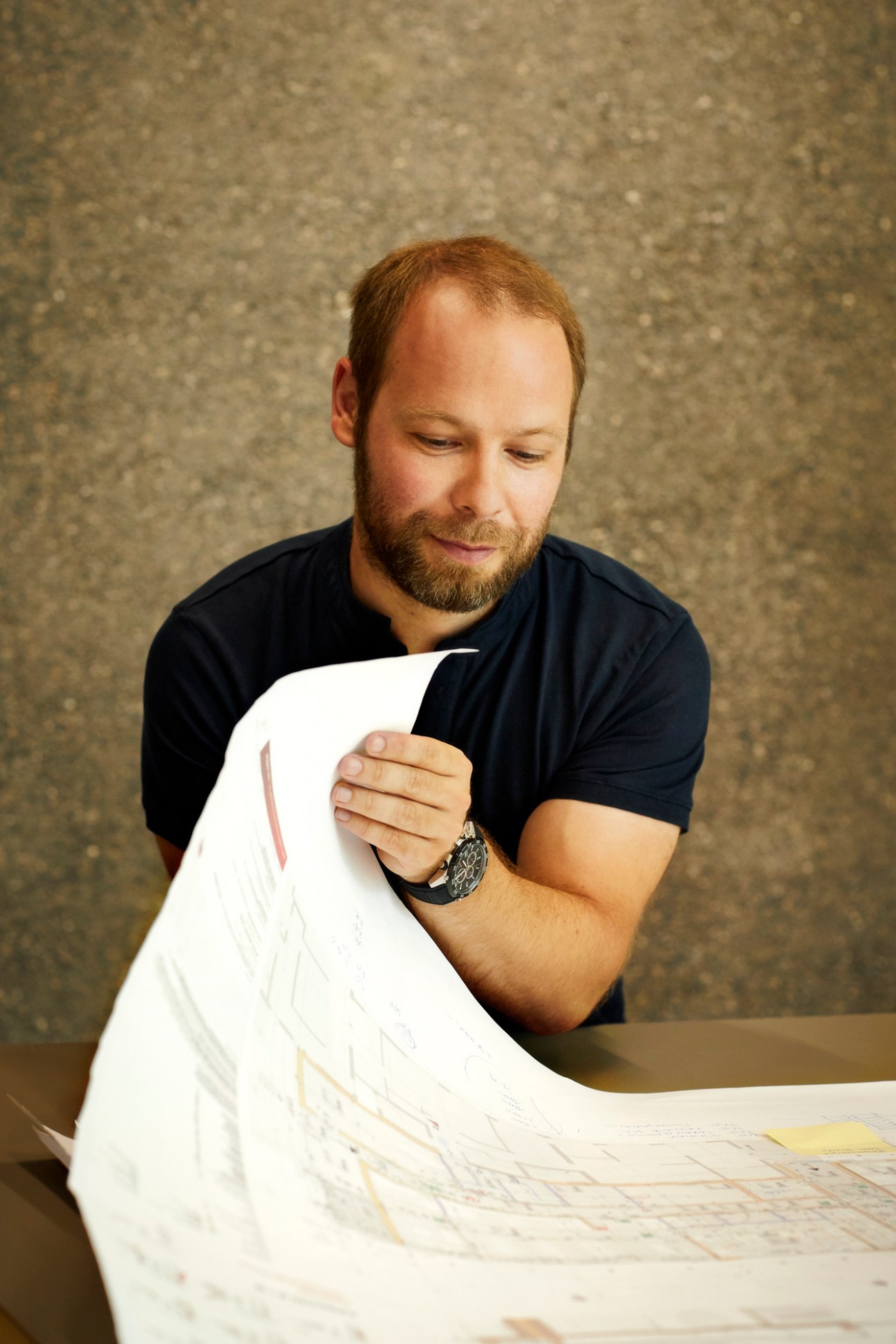Man in dark shirt examining architectural plans or documents at a workbench in a workshop setting