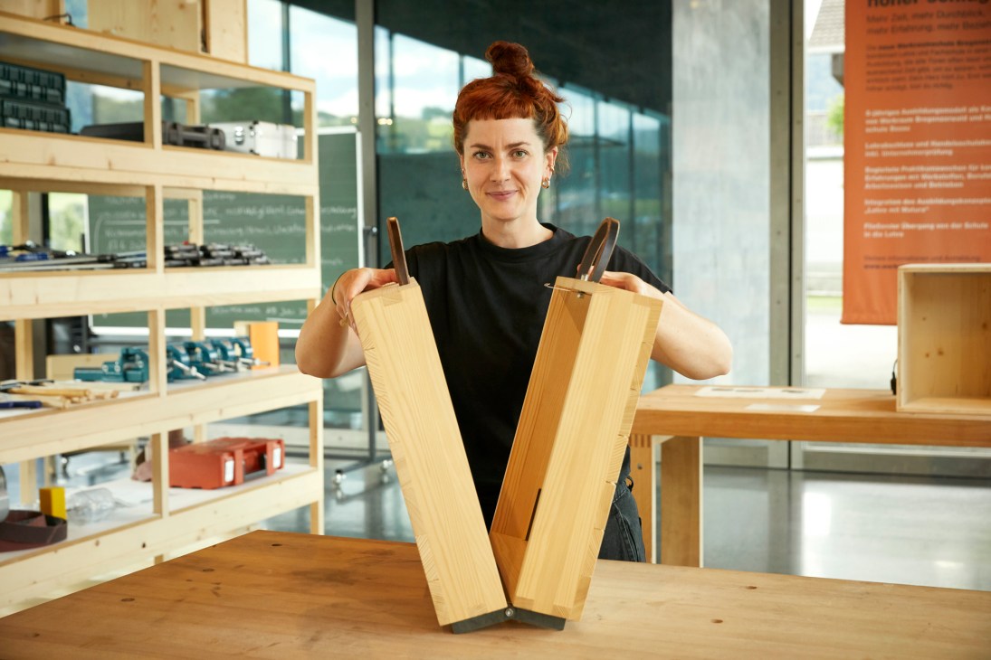 Katharina Kleiter holding wooden furniture components in a modern workshop with shelving and natural light