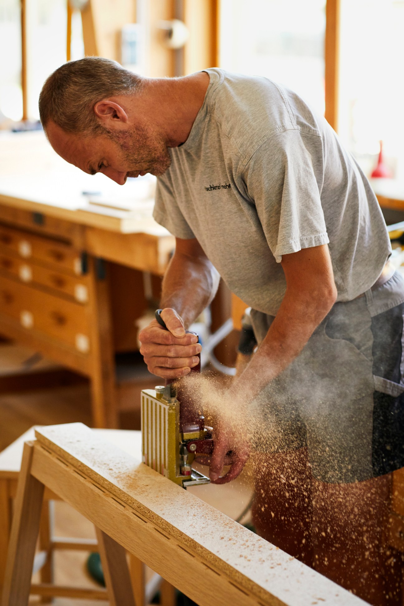 Craftsman using hand plane on wood beam creating sawdust cloud in bright timber workshop