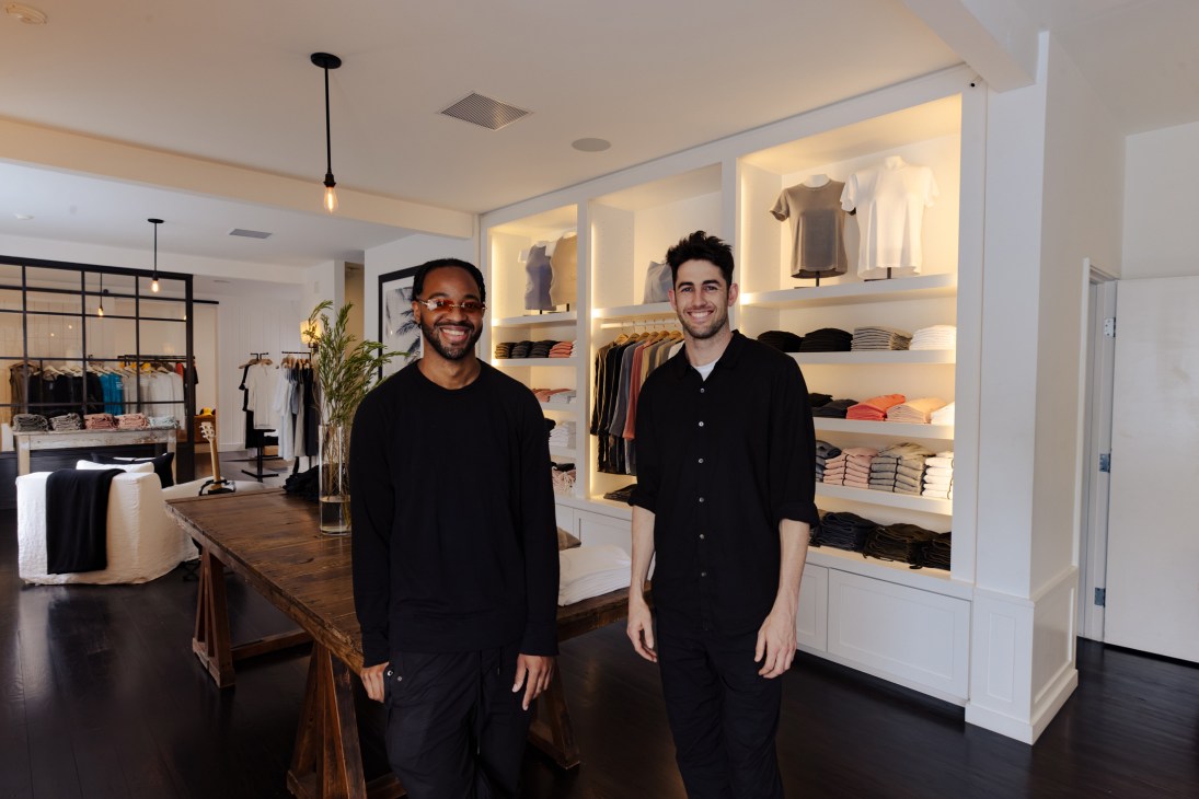Two smiling men in black clothing standing in a modern retail store with white shelving displaying folded clothes