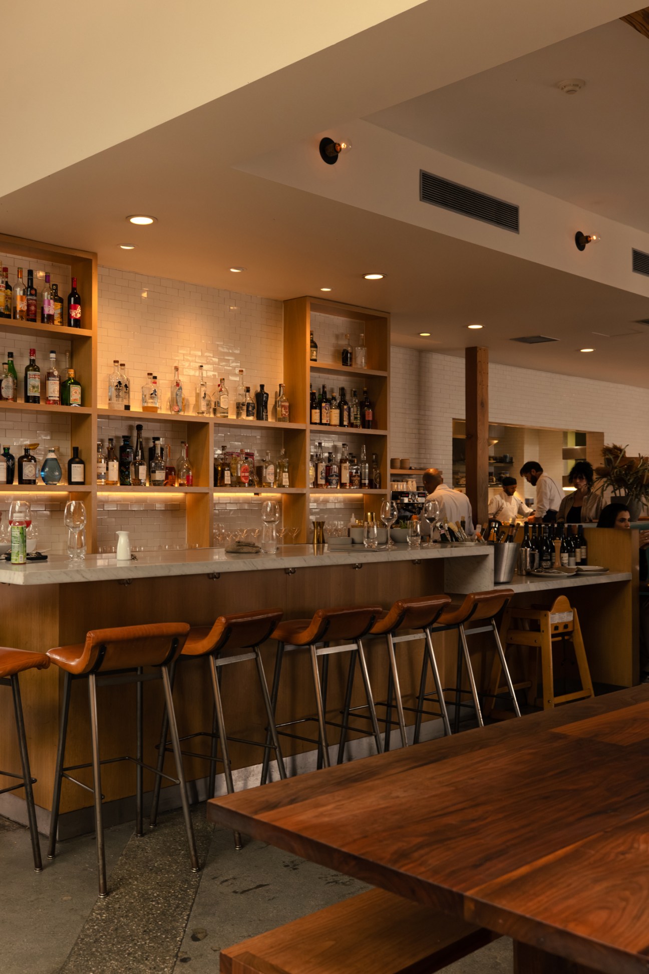 Modern restaurant bar with wooden shelving displaying bottles, orange bar stools, and staff working in background