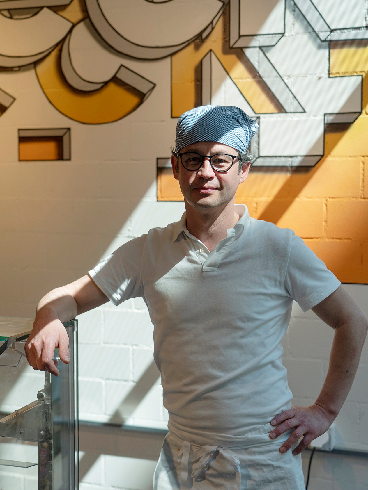 Baker Seri Wada wearing glasses and a striped cap standing in front of a colorful geometric mural