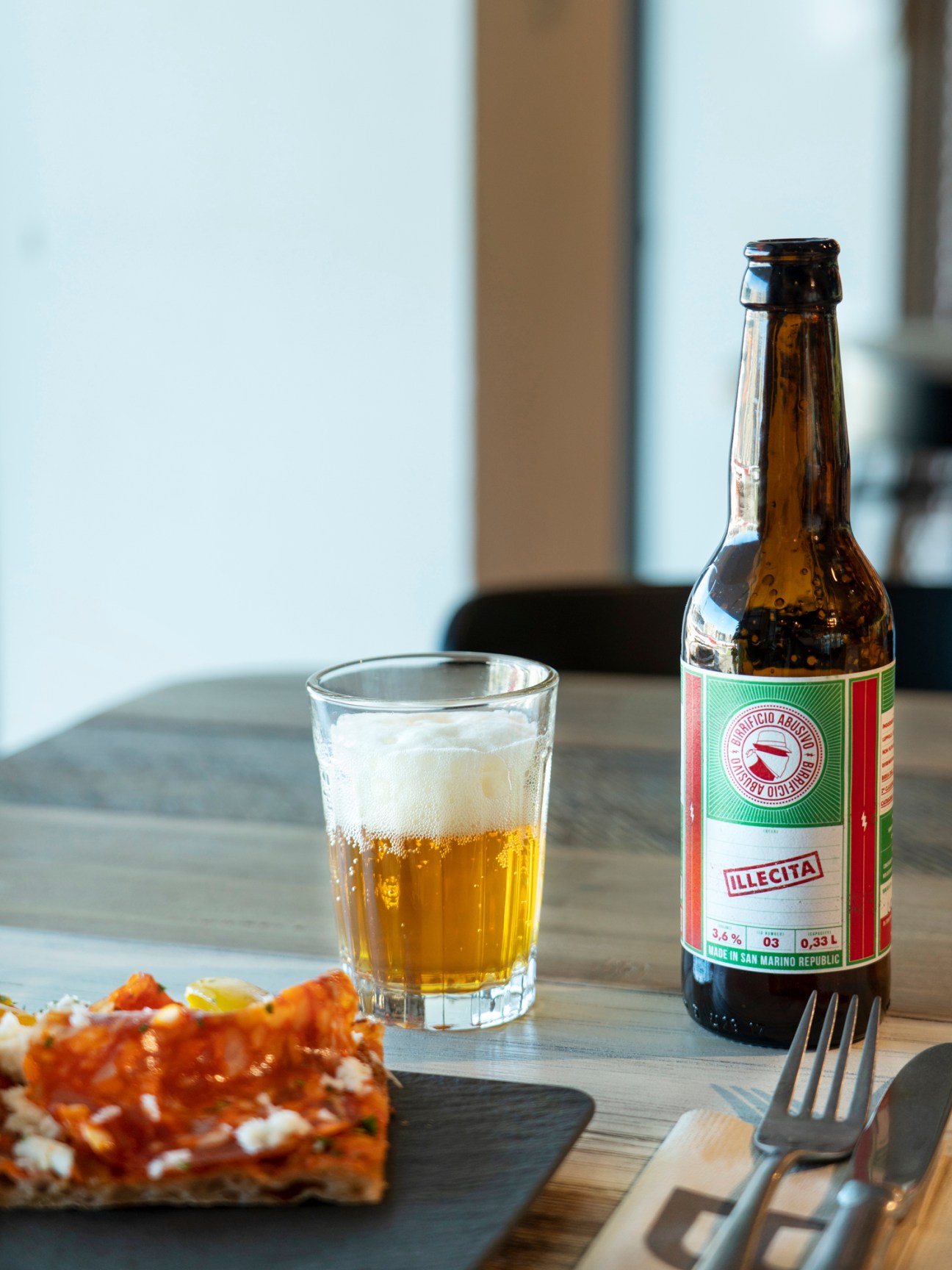 Pizza slice on slate plate with glass of beer and bottle on wooden table with cutlery