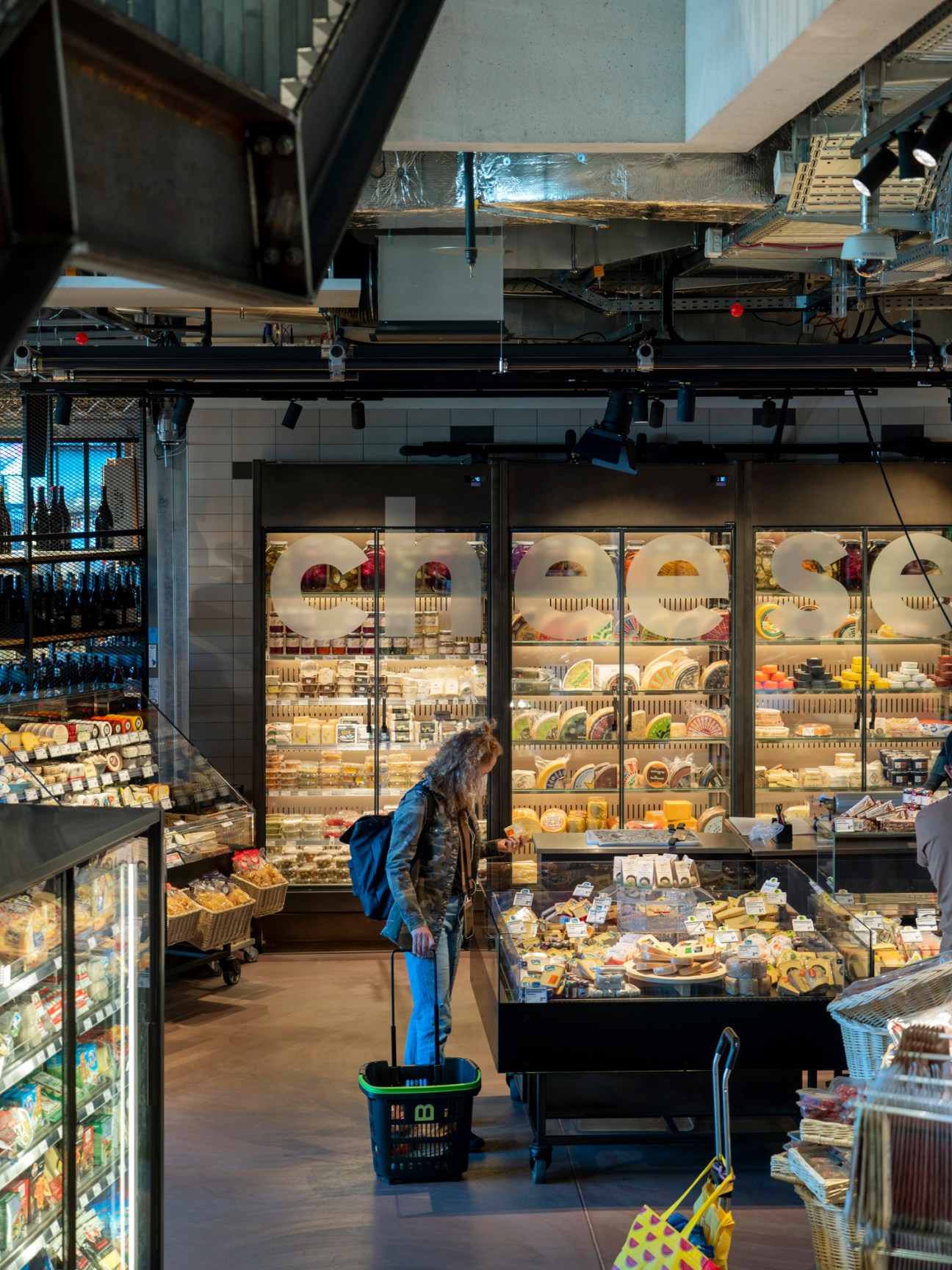 Person with shopping basket browsing large refrigerated cheese display cases in modern supermarket