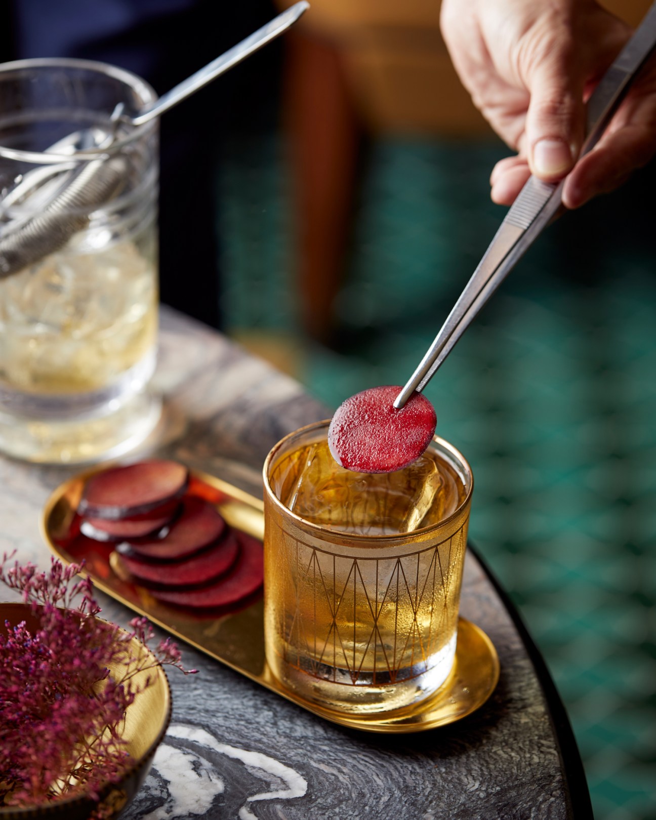 Hand using tweezers to place red beetroot disc garnish on cocktail in golden patterned glass