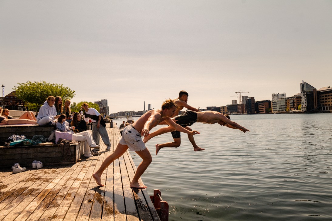 Young people jumping from a wooden dock into water with urban buildings in the background