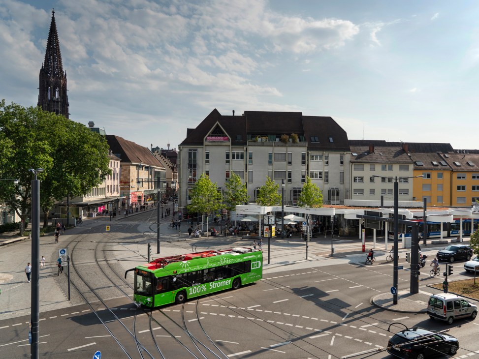 Green electric bus crossing tram tracks in Freiburg's Europaplatz with Gothic church spire and mixed-use buildings