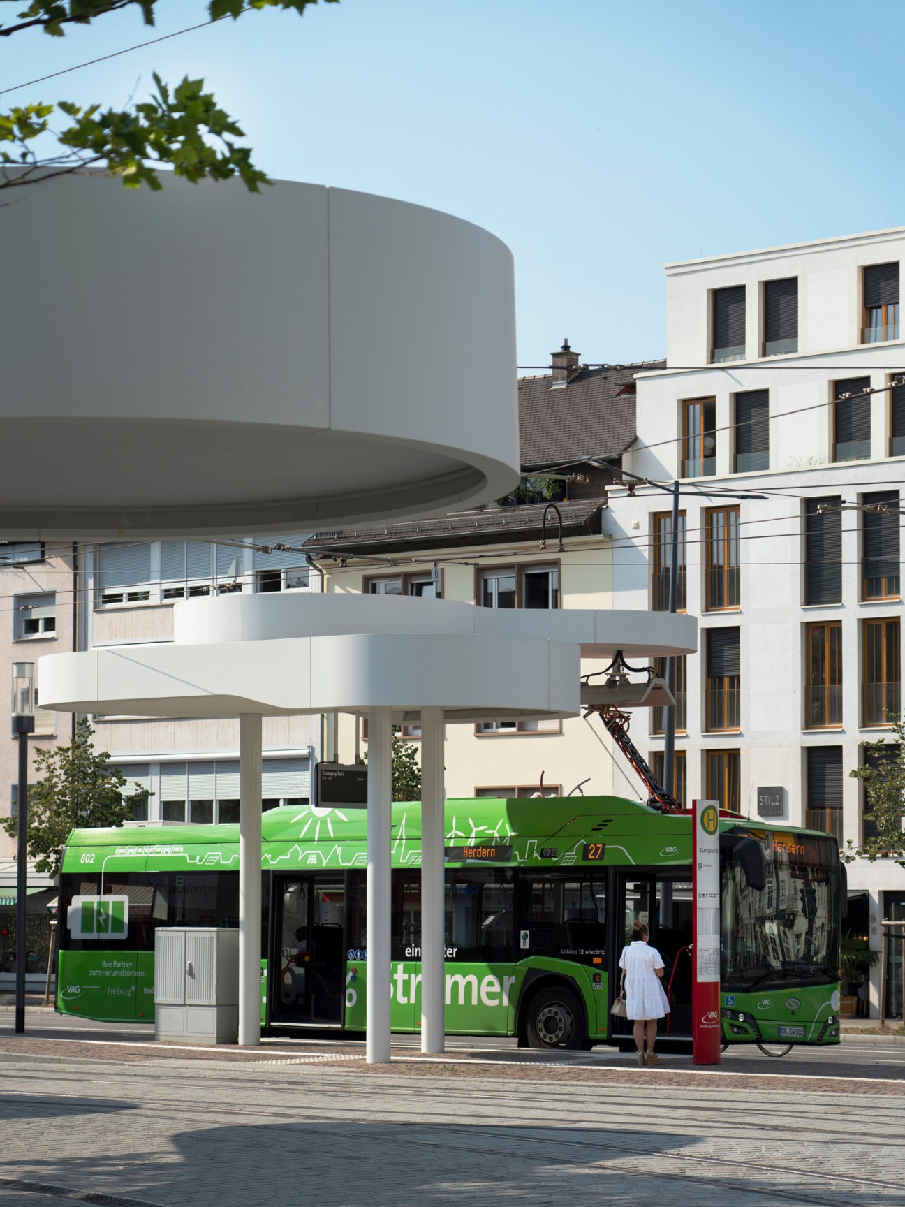 Green electric bus at modern bus stop with curved white canopy structure in urban setting