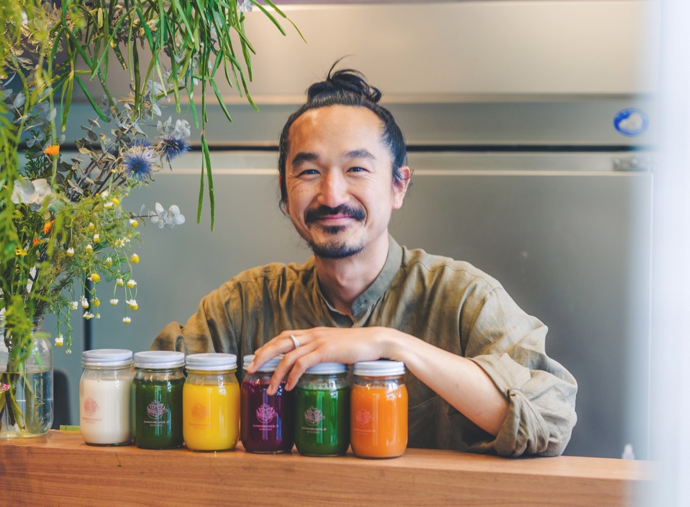 Nori Ko smiling behind a counter with colorful juice bottles in glass jars at his Tokyo shop