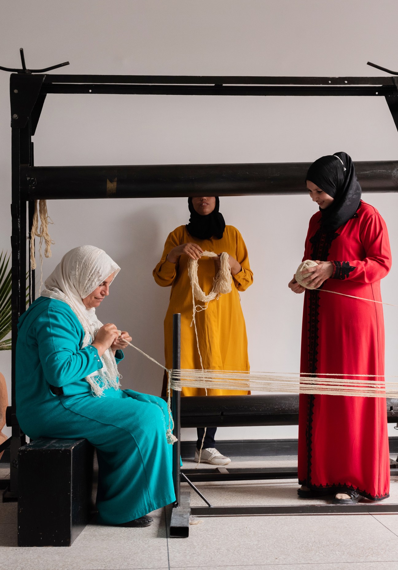 Three women in colorful traditional dress working with white threads on a large wooden loom