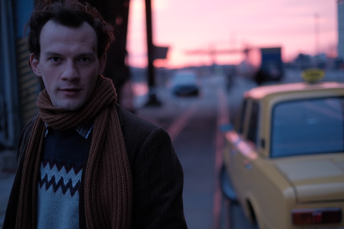 A man in dark clothing and brown knit scarf standing on a dimly lit street at dusk