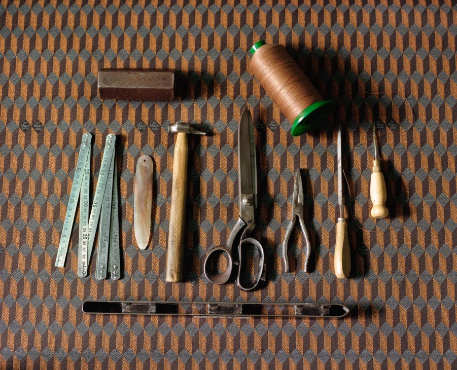 Traditional leatherworking tools including rulers, hammer, scissors, awl and thread spool arranged on patterned surface