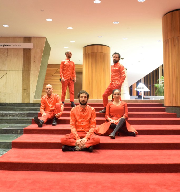 Five people in matching orange outfits sitting and standing on red carpeted stairs in a modern building lobby