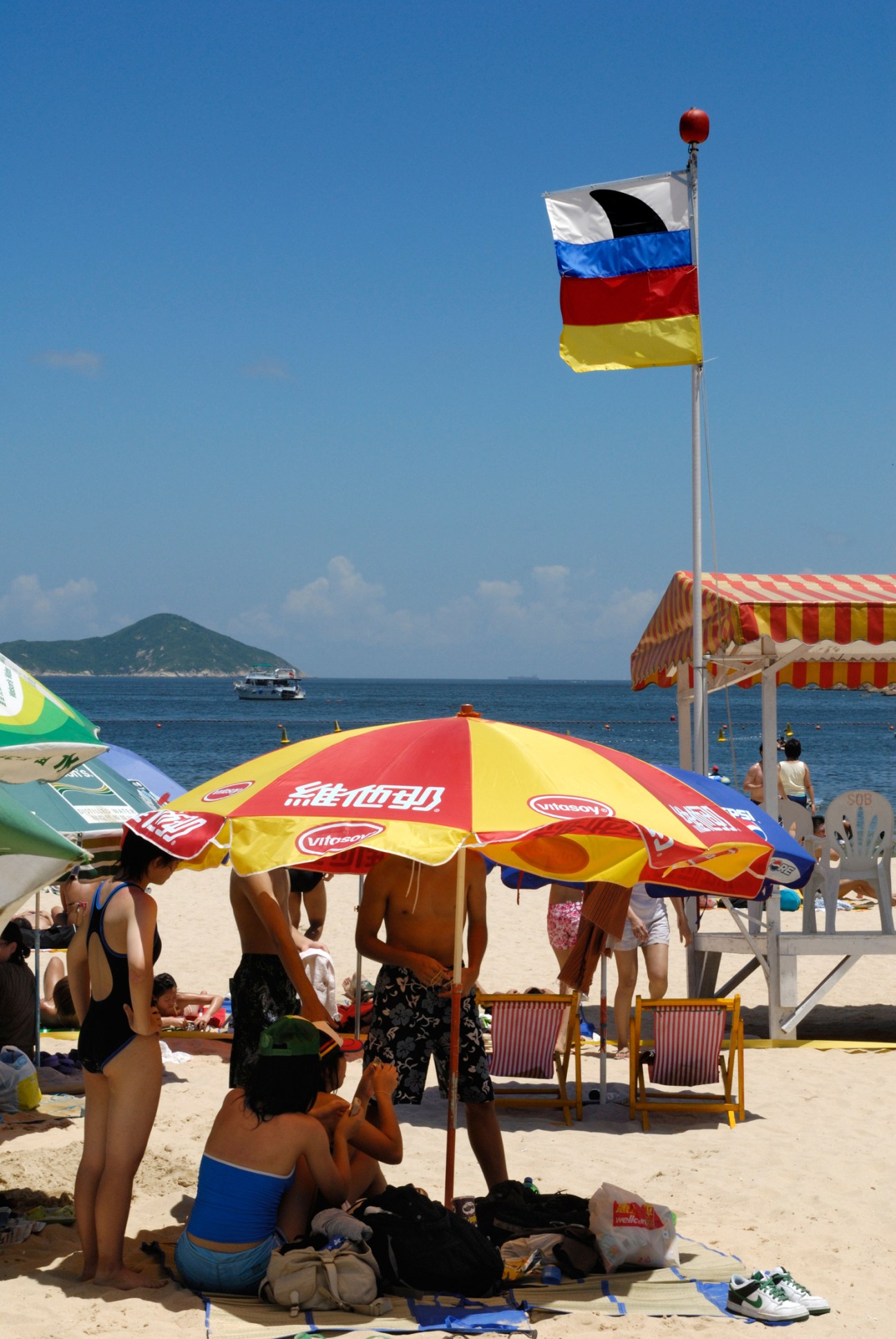 Beachgoers under colorful umbrellas on Hong Kong beach with safety flags and distant island view