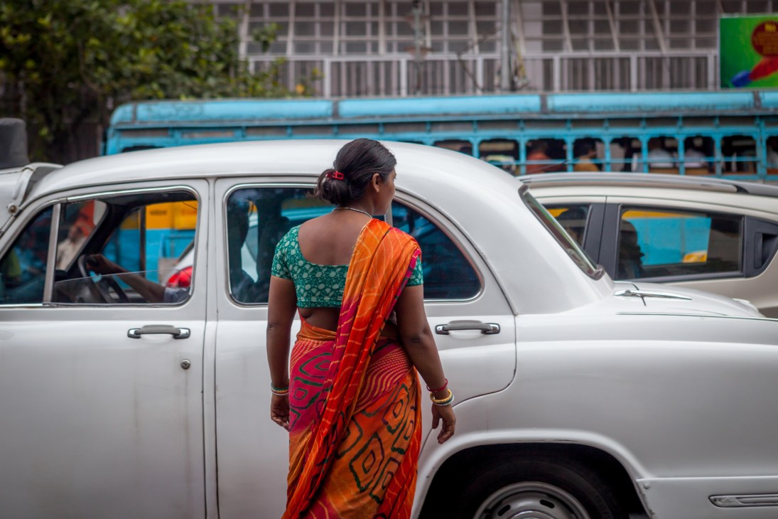 Woman in orange sari walking past white vintage car on busy street with blue bus in background