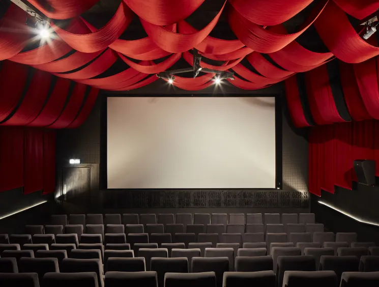 Interior of an empty cinema with dramatic red draped ceiling, grey seating, and a blank white screen.