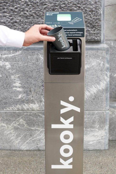 A hand deposits a dark Kooky reusable coffee cup into a grey drop-off box on a Zurich street.