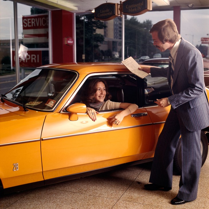 Man in pinstripe suit leaning down to talk to woman in orange sports car at gas station