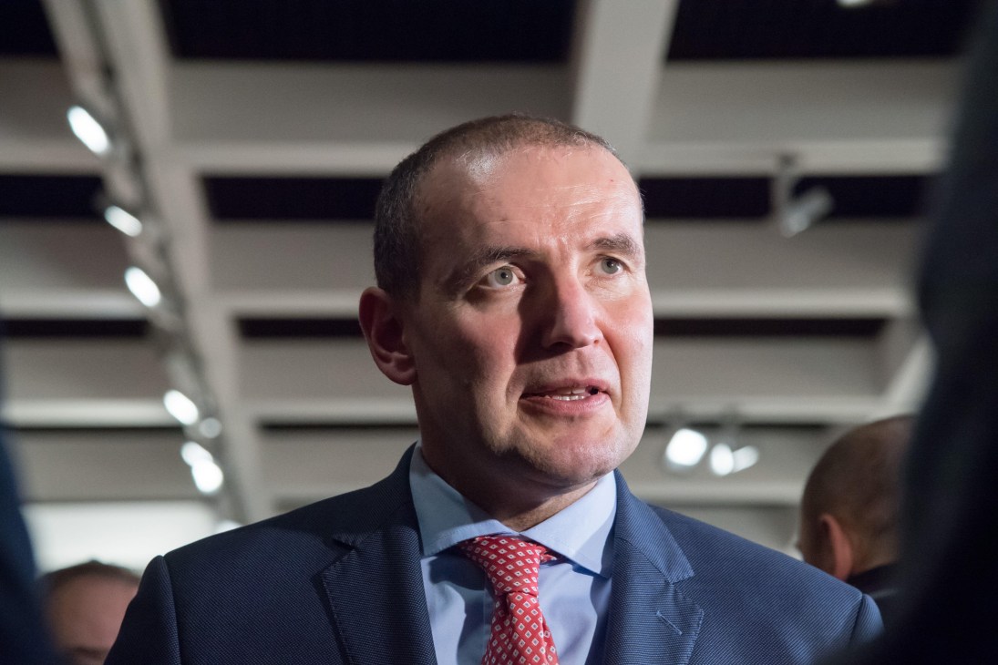 A man in a dark suit and red patterned tie speaking, photographed from a low angle under bright indoor lighting