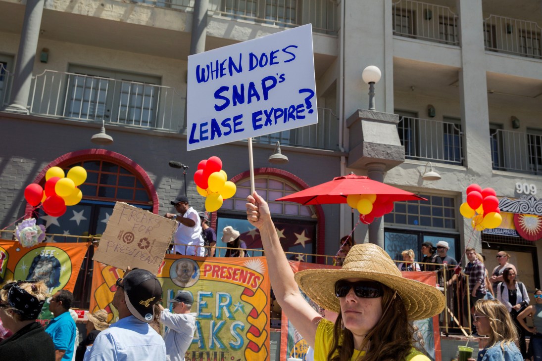 A woman wearing sunglasses and a straw hat holds up a protest sign reading 'WHEN DOES SNAP's LEASE EXPIRE?'