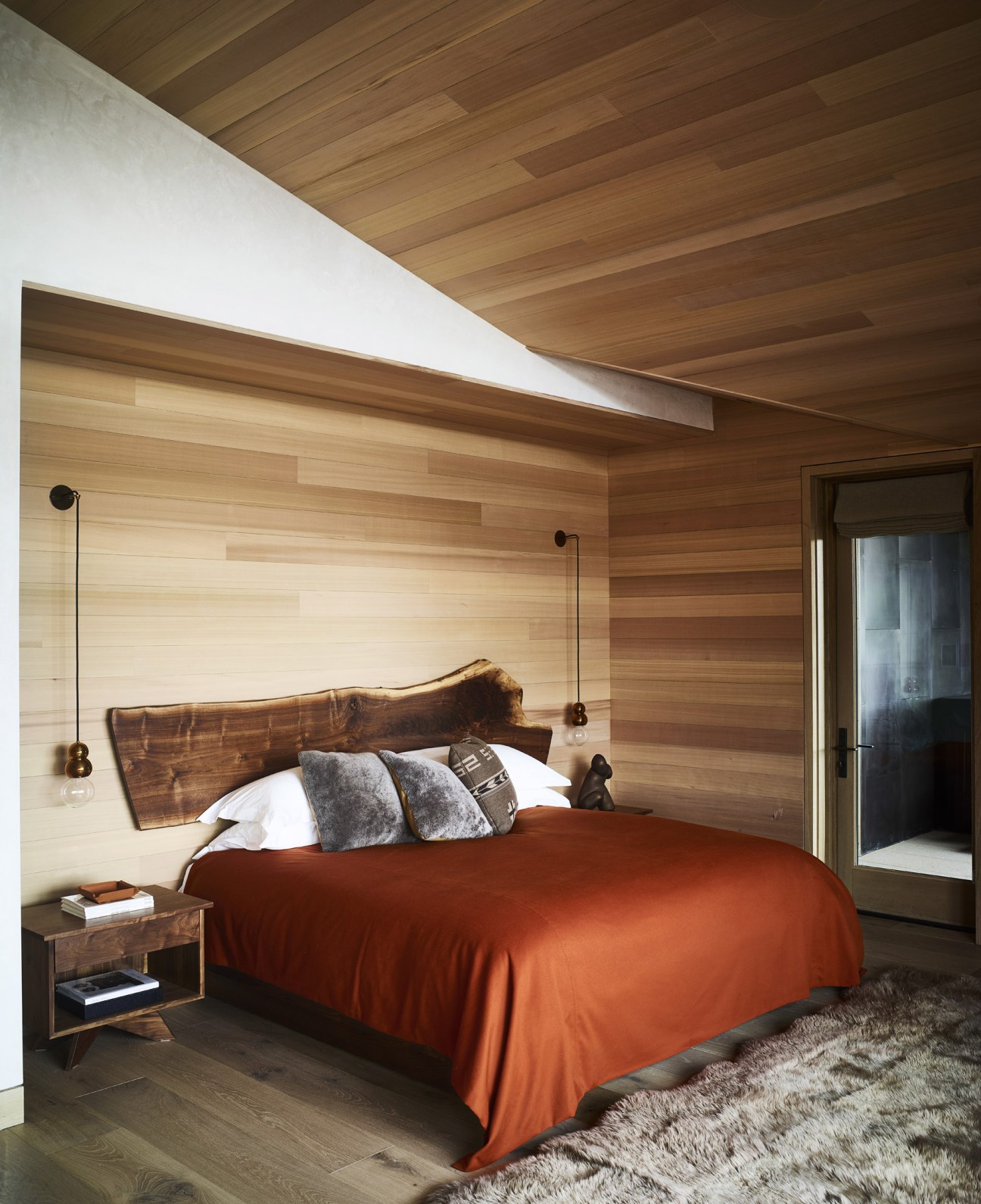 Modern bedroom with wooden ceiling, light wood-paneled walls, live-edge wooden headboard, orange bedding, pendant lights and minimalist furnishings.