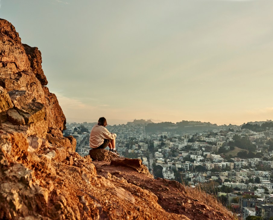 Person sitting on rocky cliff overlooking San Francisco cityscape at sunset