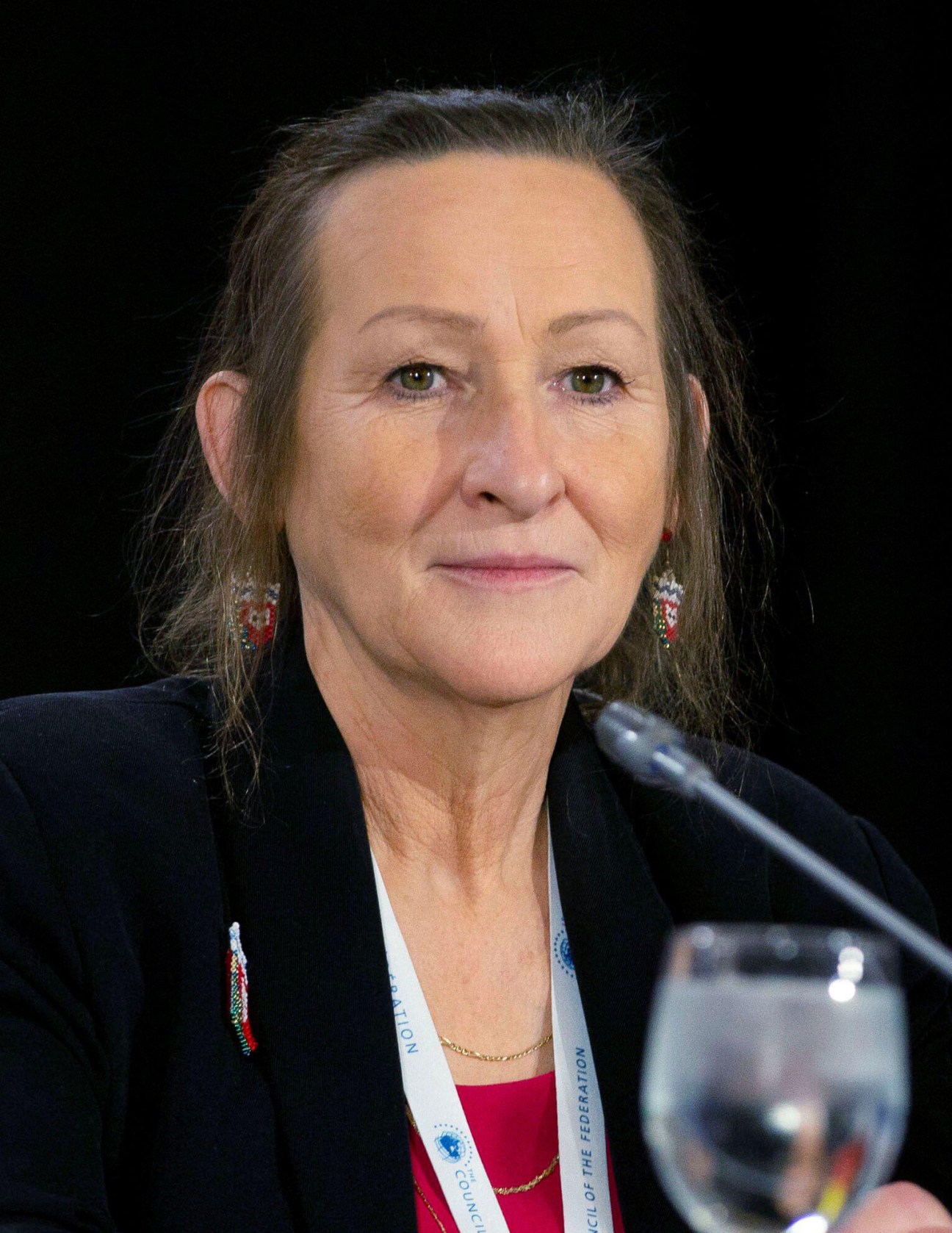 Caroline Cochrane speaking at a podium wearing a dark blazer and colorful earrings