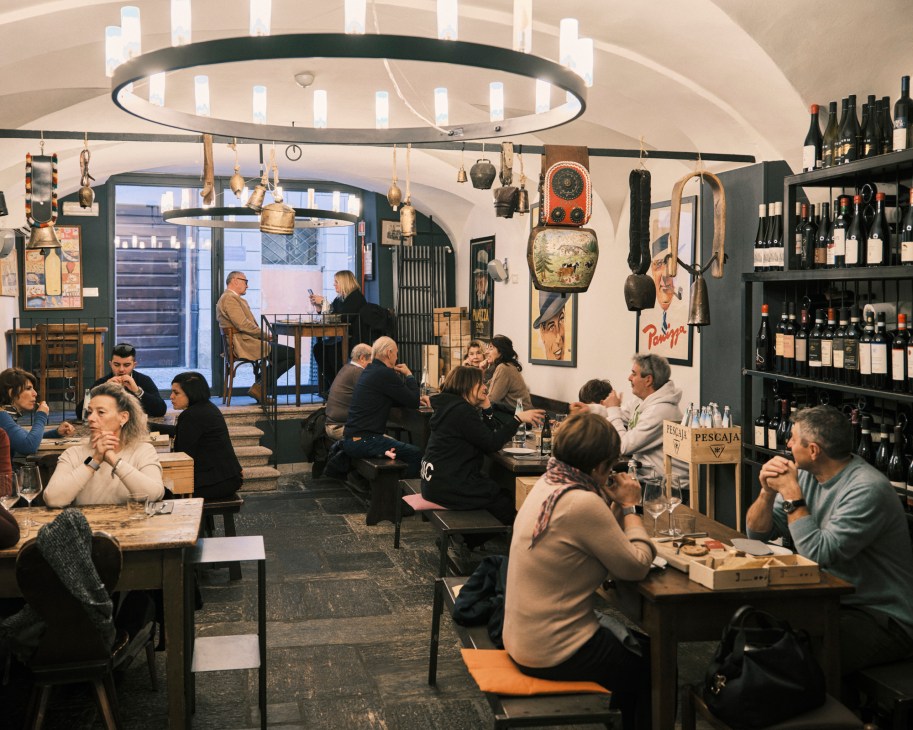 Interior of La Casera dining room with customers seated at wooden tables, wine bottles on shelves, and rustic decor