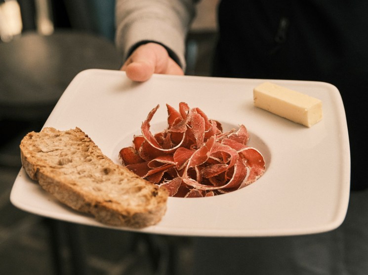 Hand holding white plate with thinly sliced cured meat, rustic bread, and cheese wedge