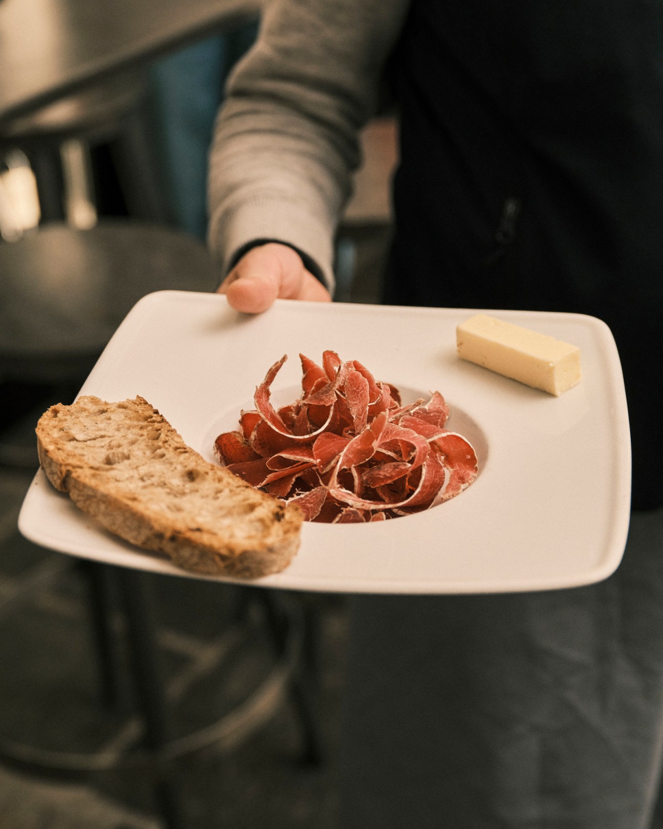 Hand holding white plate with thinly sliced cured meat, rustic bread, and cheese wedge