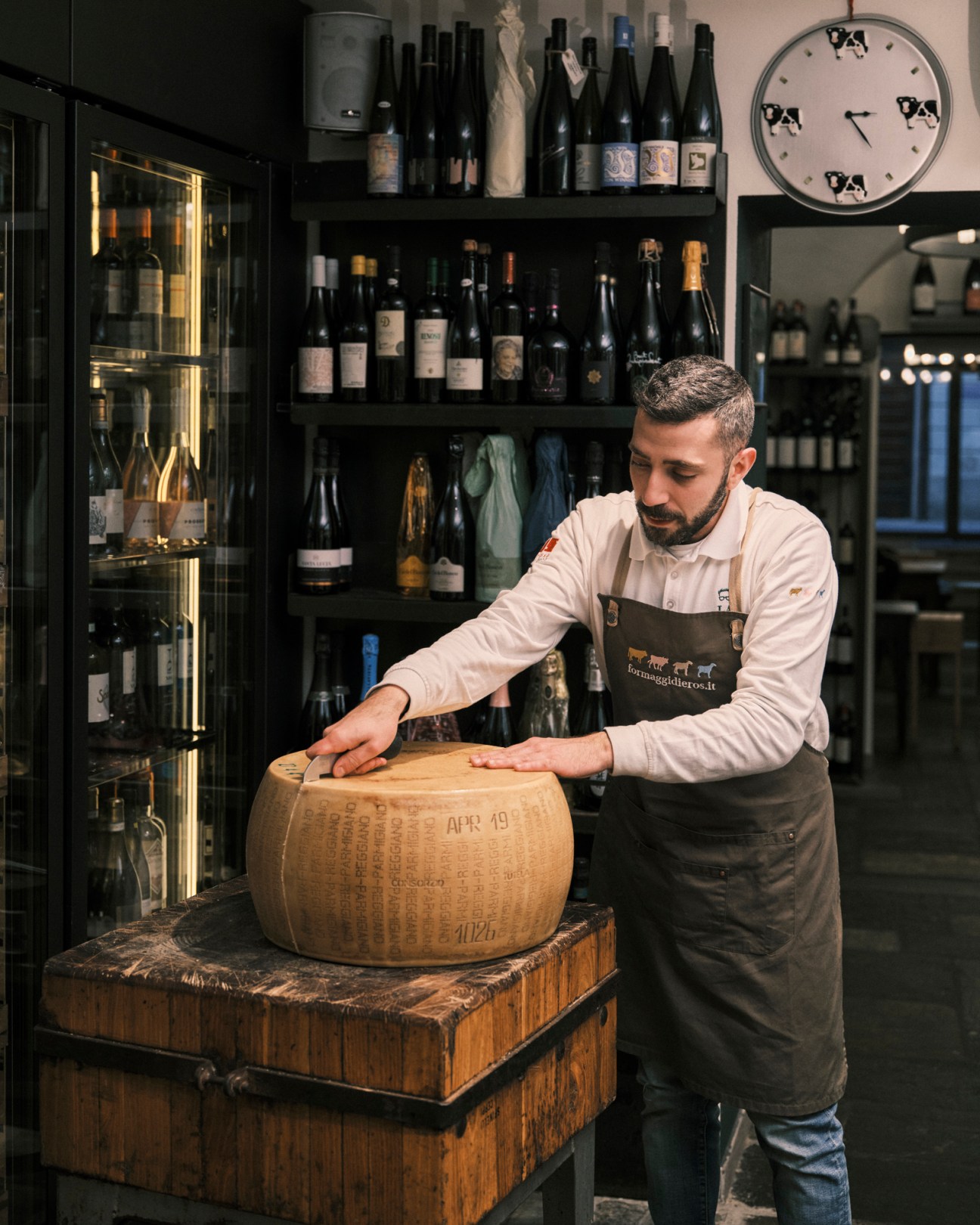 Man in apron cutting large wheel of aged cheese on wooden block in specialty food shop