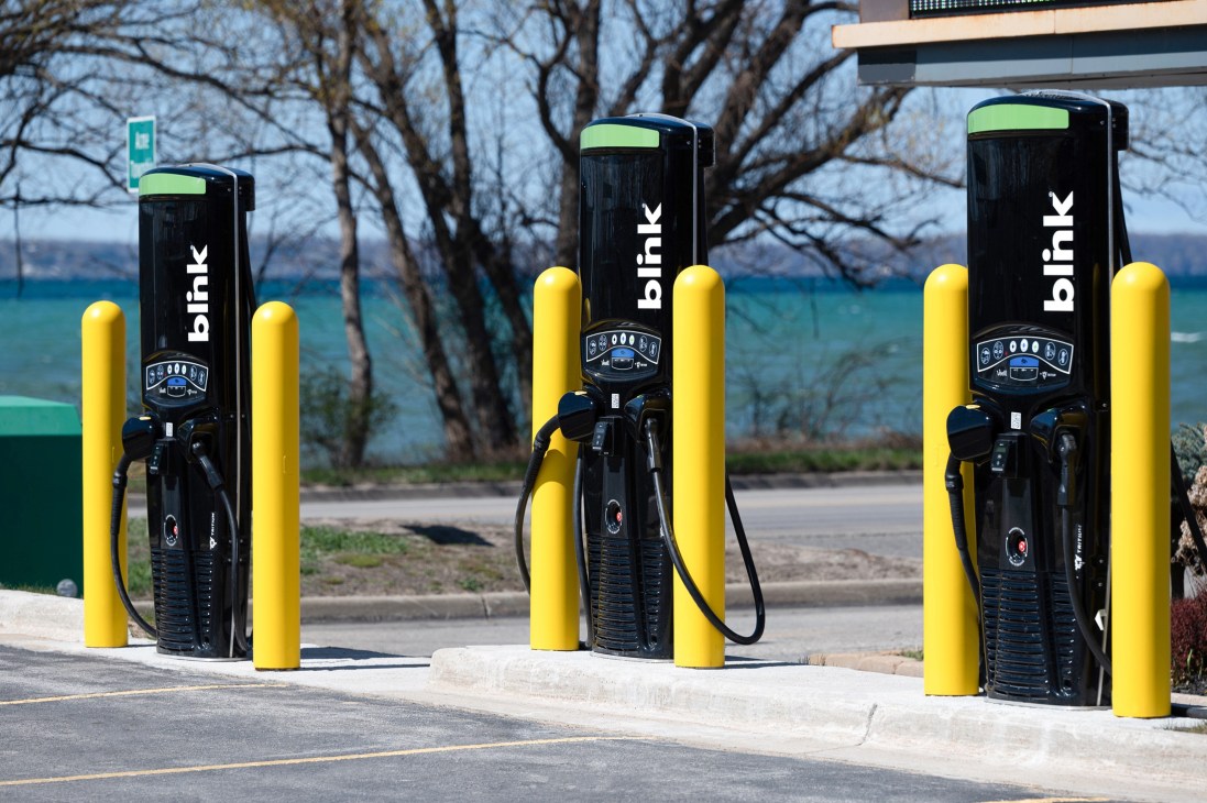Three black Blink electric vehicle charging stations with yellow protective bollards in a parking lot