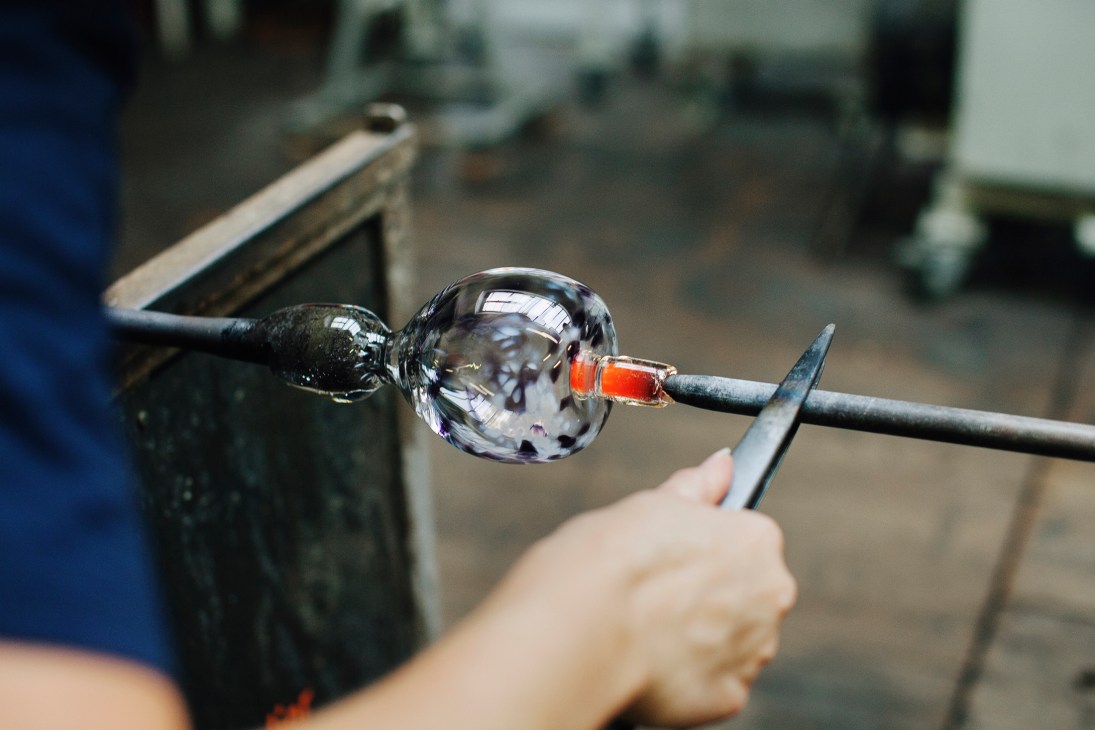 Glass artist working with molten glass on a rod, shaping heated material with tools in a workshop