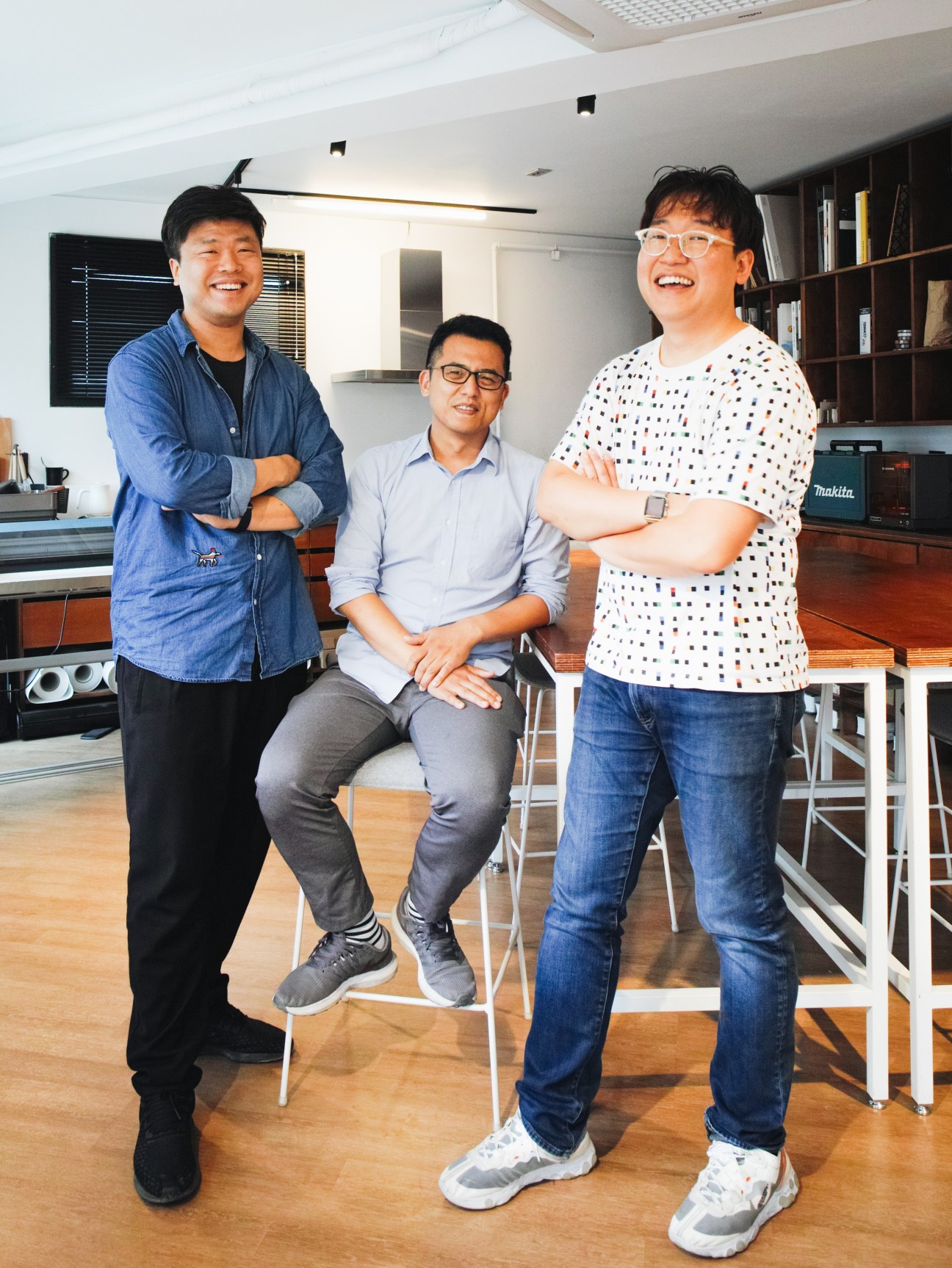 Three men smiling in a modern office space with wooden flooring and shelving.
