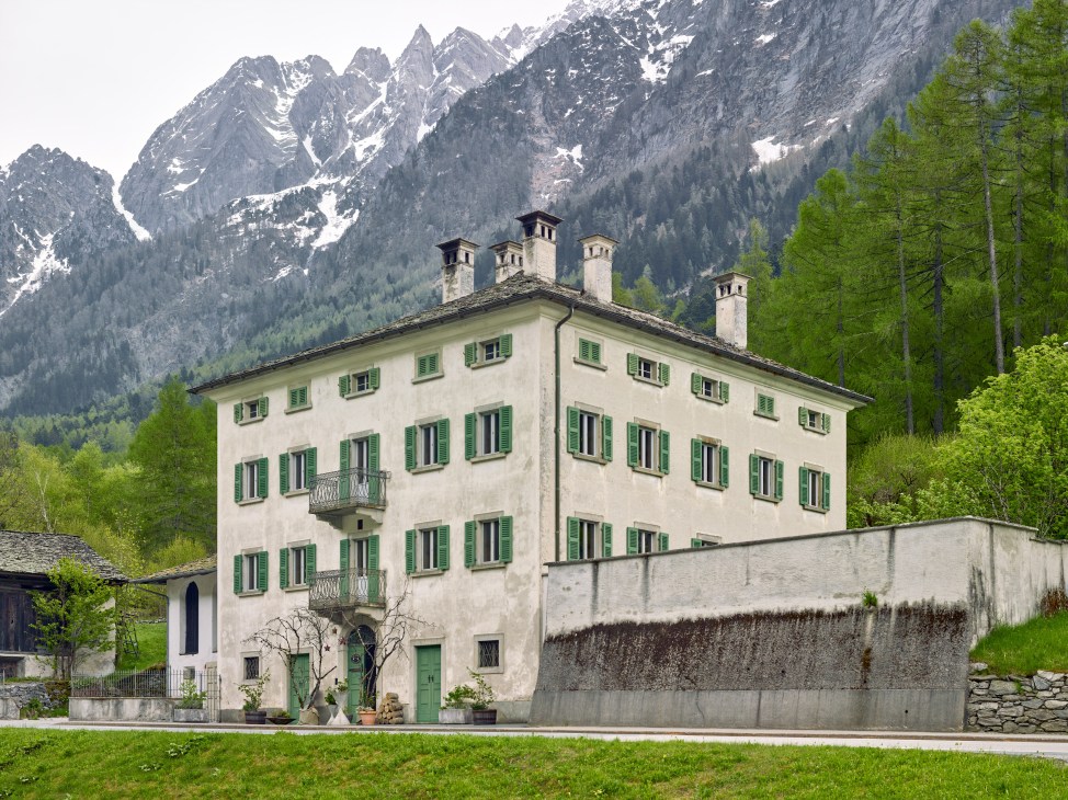 Pontisella bed and breakfast in Stampa, Switzerland, a cream-coloured historic building with green shutters set against snow-capped Alpine mountains.